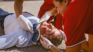 Photo of man lying on the ground with a large gash on his head, being treated by two people
